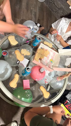 Glass-top party table with silver and green balloons, canned drinks and water, novelty sugar cookies and candy, hands arranging party favors — playful adult party scene.