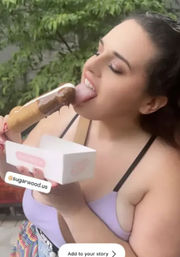 Person outdoors enjoying a chocolate-dipped popsicle while holding a white pastry box, wearing a lilac top with green trees in the background — summer dessert moment.