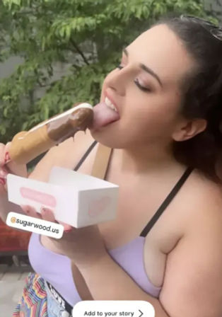 Person outdoors enjoying a chocolate-dipped popsicle while holding a white pastry box, wearing a lilac top with green trees in the background — summer dessert moment.