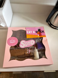 Pink pastry kit box with clear window on a white kitchen counter, playful stickers and a colorful card reading 'EAT A DICK'.