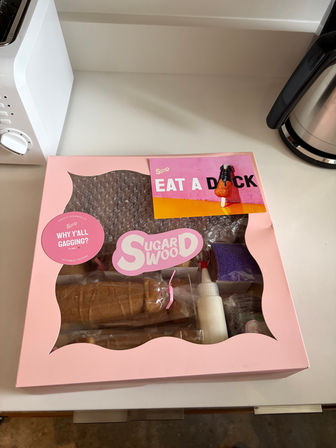Pink pastry kit box with clear window on a white kitchen counter, playful stickers and a colorful card reading 'EAT A DICK'.