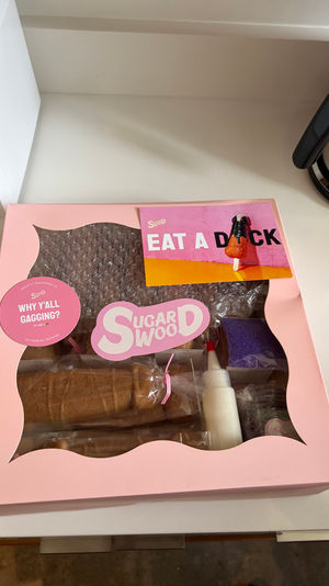 Pink pastry kit box with clear window on a white kitchen counter, playful stickers and a colorful card reading 'EAT A DICK'.