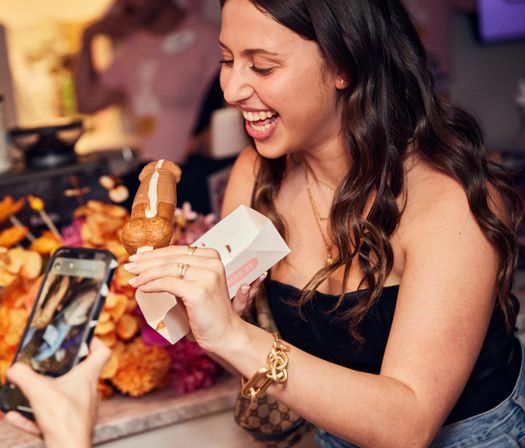 Laughing woman at an indoor event holding a chocolate‑glazed éclair in a paper tray while a friend photographs it on a smartphone, colorful dessert display behind