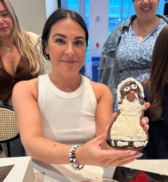 Smiling woman at a kitchen counter holding a hand-decorated gingerbread cookie shaped like a person in a white dress, friends gathered in the bright indoor party setting.