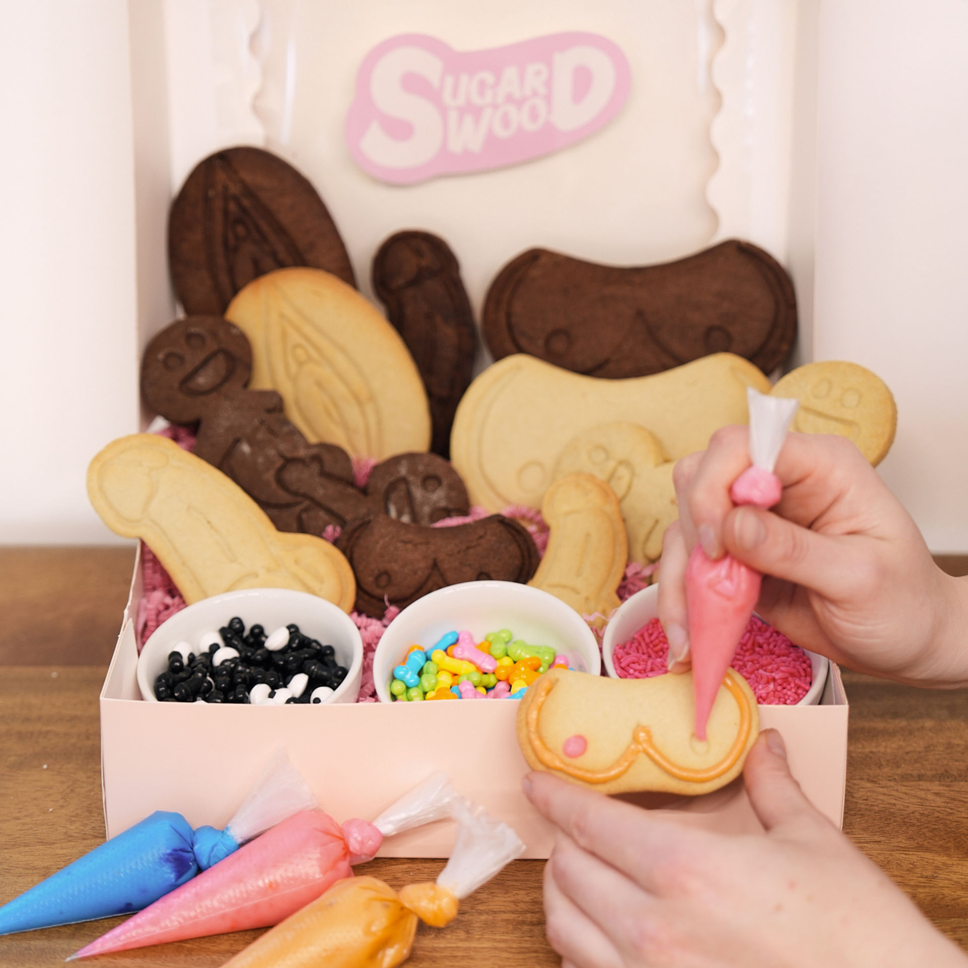 Hands piping pink icing onto a novelty cookie inside a box of chocolate and vanilla penis- and vulva-shaped cookies, with bowls of sprinkles and candy — adult novelty cookie decorating kit on a wooden table.
