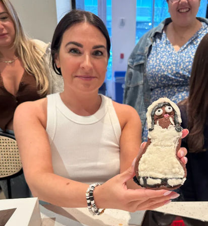 Smiling woman in a white tank top holds a whimsical frosted cookie shaped like a person in a white robe, surrounded by friends at an indoor gathering around a countertop.