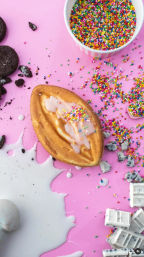Golden mini waffle with white glaze and rainbow nonpareils on a pink surface, surrounded by a bowl of multicolored sprinkles, scattered nonpareils, crumbled chocolate sandwich cookies, spilled milk and white chocolate squares.