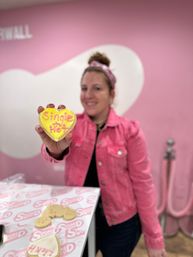 Smiling person in a pink jacket holds a yellow heart-shaped cookie iced "Single n' Hot" in a pink, heart-themed bakery with decorated cookies on the counter.