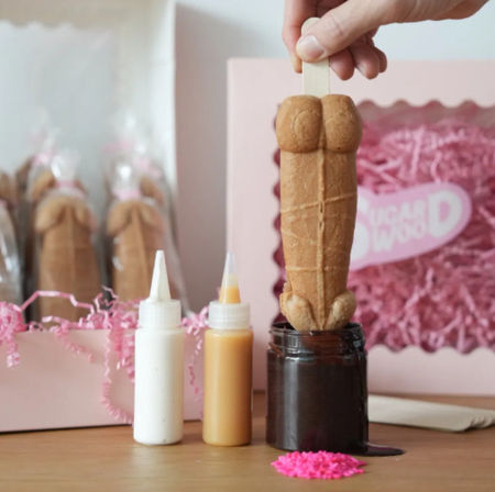 Hand holding an adult-themed novelty cookie pop on a stick being dipped in chocolate, with squeeze-icing bottles, pink gift box and pink sprinkles