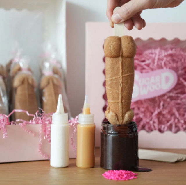 Hand holding an adult-themed novelty cookie pop on a stick being dipped in chocolate, with squeeze-icing bottles, pink gift box and pink sprinkles