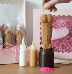 Hand holding a playful body-shaped cookie pop on a stick being dipped into chocolate sauce, with squeeze bottles of white and caramel drizzle, pink sprinkles, and a pink gift box with shredded paper in the background — dessert gift presentation.