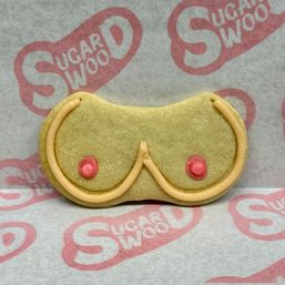 Novelty boob-shaped sugar cookie decorated with piped tan icing and pink icing dots, photographed on branded parchment — playful bakery-style treat
