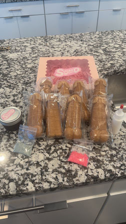 Playful adult cookie kit on speckled granite kitchen countertop: individually wrapped penis-shaped cookies, pink bakery box, small bottle of icing and packets of pink and silver sprinkles.