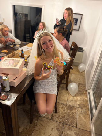 Smiling bride-to-be in a sequin dress and 'BRIDE' headband holding decorated cookies at a cozy home dining-table bachelorette party with friends, pastry boxes and white balloons.