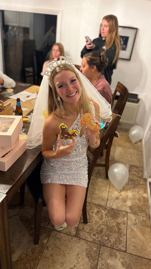 Smiling bride-to-be in a sequin dress and 'BRIDE' headband holding decorated cookies at a cozy home dining-table bachelorette party with friends, pastry boxes and white balloons.
