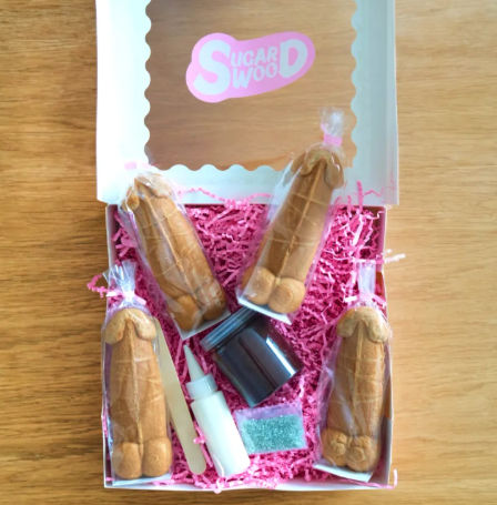 White gift box on a wooden table holding four individually wrapped novelty penis-shaped cookies, icing bottle, sprinkles and pink paper shred for decorating