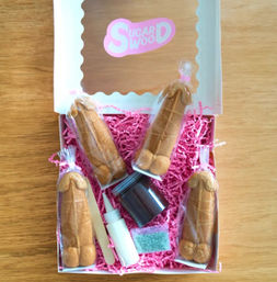 White gift box on a wooden table holding four individually wrapped novelty penis-shaped cookies, icing bottle, sprinkles and pink paper shred for decorating