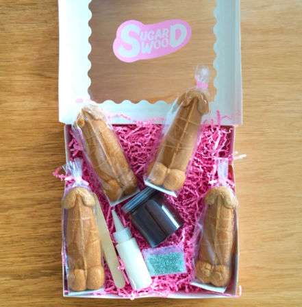 White gift box on a wooden table holding four individually wrapped novelty penis-shaped cookies, icing bottle, sprinkles and pink paper shred for decorating