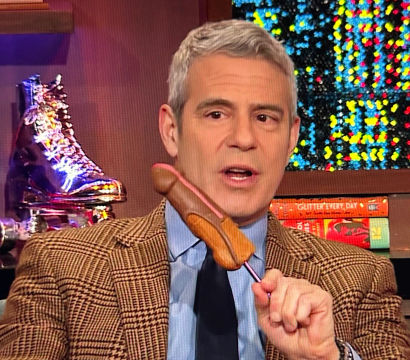 TV host in a brown plaid blazer on a colorful television studio set holding a corn dog on a pink stick, glittery backdrop and metallic roller skate prop visible behind him