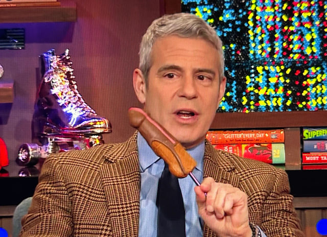 TV host in a brown plaid blazer on a colorful television studio set holding a corn dog on a pink stick, glittery backdrop and metallic roller skate prop visible behind him