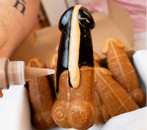 Close-up of a novelty adult-shaped waffle pastry being drizzled with chocolate glaze and caramel sauce in a bakery box — playful dessert treat