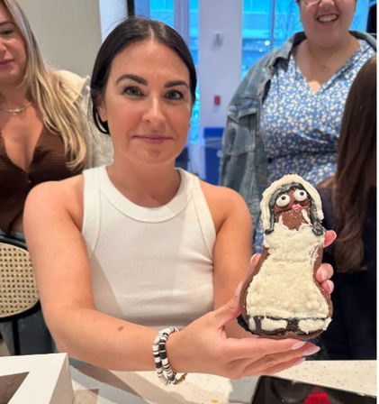 Smiling woman in a white tank top holding a large decorated gingerbread cookie shaped like a person with white icing dress, friends laughing behind her in a bright indoor cafe or kitchen setting.