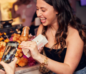 Laughing woman holding a glazed éclair in a small takeout box while someone photographs it with a smartphone, colorful flowers blurred in the background.