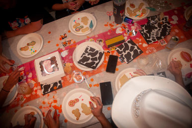 Top-down view of a lively cookie-decorating party: hands icing gingerbread and shaped cookies on paper plates, cow-print napkins, orange confetti, phones and drinks scattered on the table.