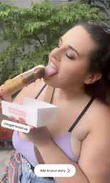Person enjoying a chocolate-dipped ice cream bar on a stick outdoors on a leafy patio, smiling while holding a takeaway box — fun summer dessert moment.