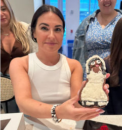 Smiling woman in a white tank top at an indoor cafe holding a large hand-decorated iced cookie shaped like a cartoon person with white frosting dress and candy eyes, friends behind her.