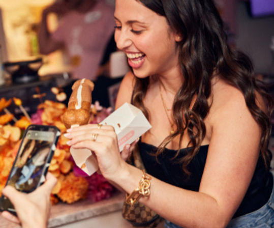 Smiling woman at an indoor party holding a chocolate-glazed cream-filled pastry on a stick in a takeaway box while a friend photographs it with a smartphone, colorful flowers in the background.