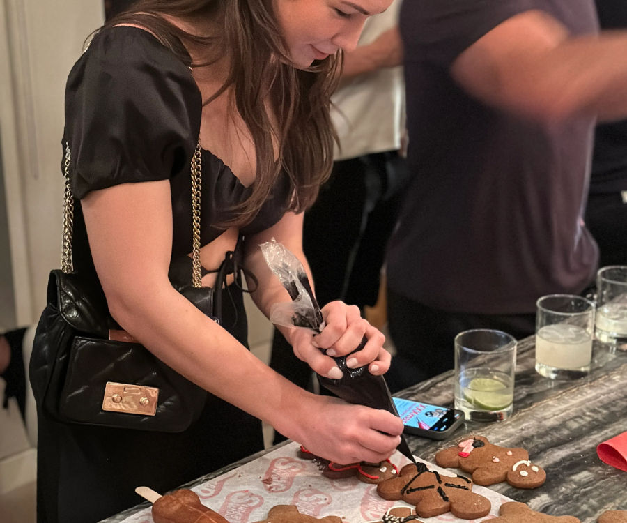 Young woman piping black icing onto gingerbread cookies on a kitchen counter at a festive gathering, with cocktail glasses and a smartphone nearby