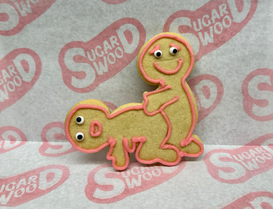 Novelty decorated sugar cookie shaped like two cartoon figures in a playful pose, tan cookie outlined with pink icing, candy eyes and smiles, sitting on patterned parchment paper.