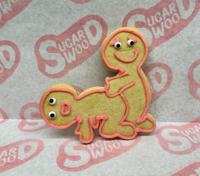 Playful suggestive sugar cookie shaped as two gingerbread figures in a cheeky pose, outlined with pink icing and candy eyes on parchment paper.