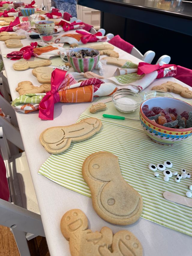 Bright kitchen table set for a cookie-decorating party with striped placemats, colorful bowls of gummy candies and sugar, undecorated sugar cookies in various shapes (gingerbread and mask-like), and napkins tied with pink ribbons.