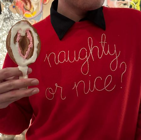 Close-up of a person in a red holiday sweater embroidered “naughty or nice?” holding a spoon-shaped frosted cookie with pink and white icing, playful festive scene.