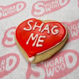 Heart-shaped Valentine sugar cookie with glossy red icing and white piped "SHAG ME" message, bakery-style on pink patterned parchment paper