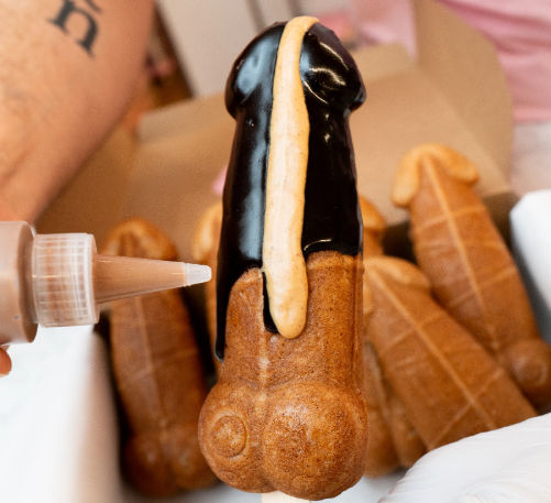Close-up of a novelty penis-shaped waffle on a stick being drizzled with chocolate and caramel sauce, with additional similar pastries in the background
