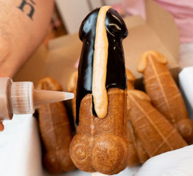 Close-up of a novelty penis-shaped waffle on a stick being drizzled with chocolate and caramel sauce, with additional similar pastries in the background