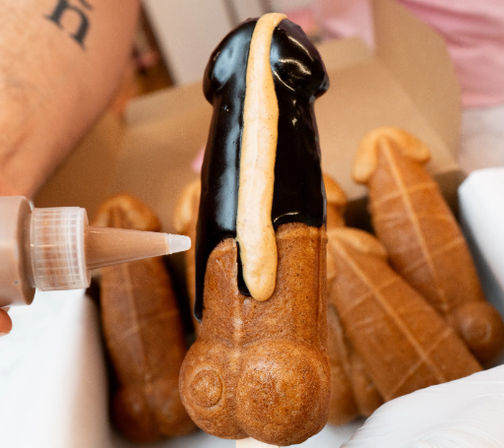 Playful adult-themed novelty pastry shaped like a penis, close-up as chocolate glaze and caramel drizzle are piped over it in a pastry box