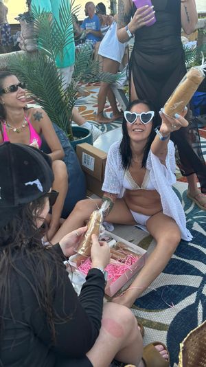 Sunny beachside bachelorette party with women in swimsuits and heart-shaped sunglasses laughing on a patterned rug, exchanging wrapped novelty treats amid tropical potted plants.