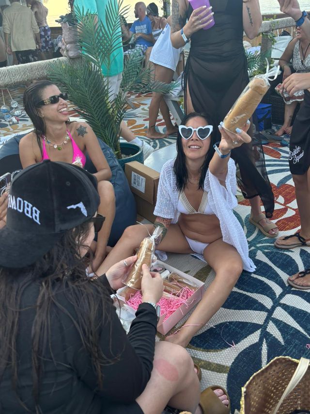 Sunny beachside bachelorette party with women in swimsuits and heart-shaped sunglasses laughing on a patterned rug, exchanging wrapped novelty treats amid tropical potted plants.