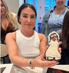 Woman in a white tank top holding a decorated gingerbread cookie shaped like a bride, friends behind her at an indoor gathering