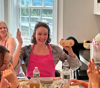 Smiling woman in a pink apron holds up a decorated cookie while friends applaud at a lively cookie-decorating party in a sunlit home kitchen, table scattered with iced cookies, bowls and drinks.