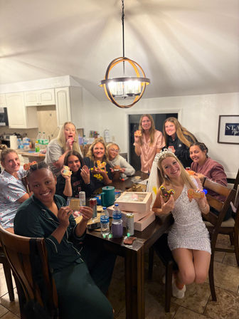 Smiling friends gathered in a home kitchen dining area for a bachelorette cookie-decorating party, bride-to-be in a veil holding decorated cookies at a table with pastry boxes and a modern pendant light