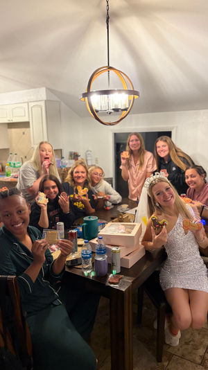 Smiling friends gathered in a home kitchen dining area for a bachelorette cookie-decorating party, bride-to-be in a veil holding decorated cookies at a table with pastry boxes and a modern pendant light