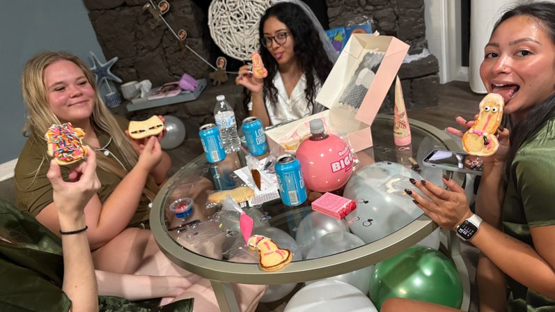 Three friends at an indoor bachelorette-style party around a glass table, holding and eating decorated novelty cookies with balloons, drinks and a cookie box nearby.