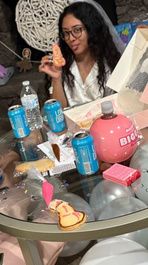 Three friends at an indoor bachelorette-style party around a glass table, holding and eating decorated novelty cookies with balloons, drinks and a cookie box nearby.
