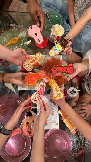 At-home living room cookie-decorating party with friends’ hands holding colorful, decorated penis-shaped cookies over a glass coffee table with frosting tubs, piping bags and pink bowls.