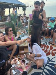 Group of friends at a beachfront tiki-style bar laughing and sharing novelty pastries, woman in heart-shaped sunglasses kneeling as others hand a pastry, colorful patterned rug and ocean sunset in the background